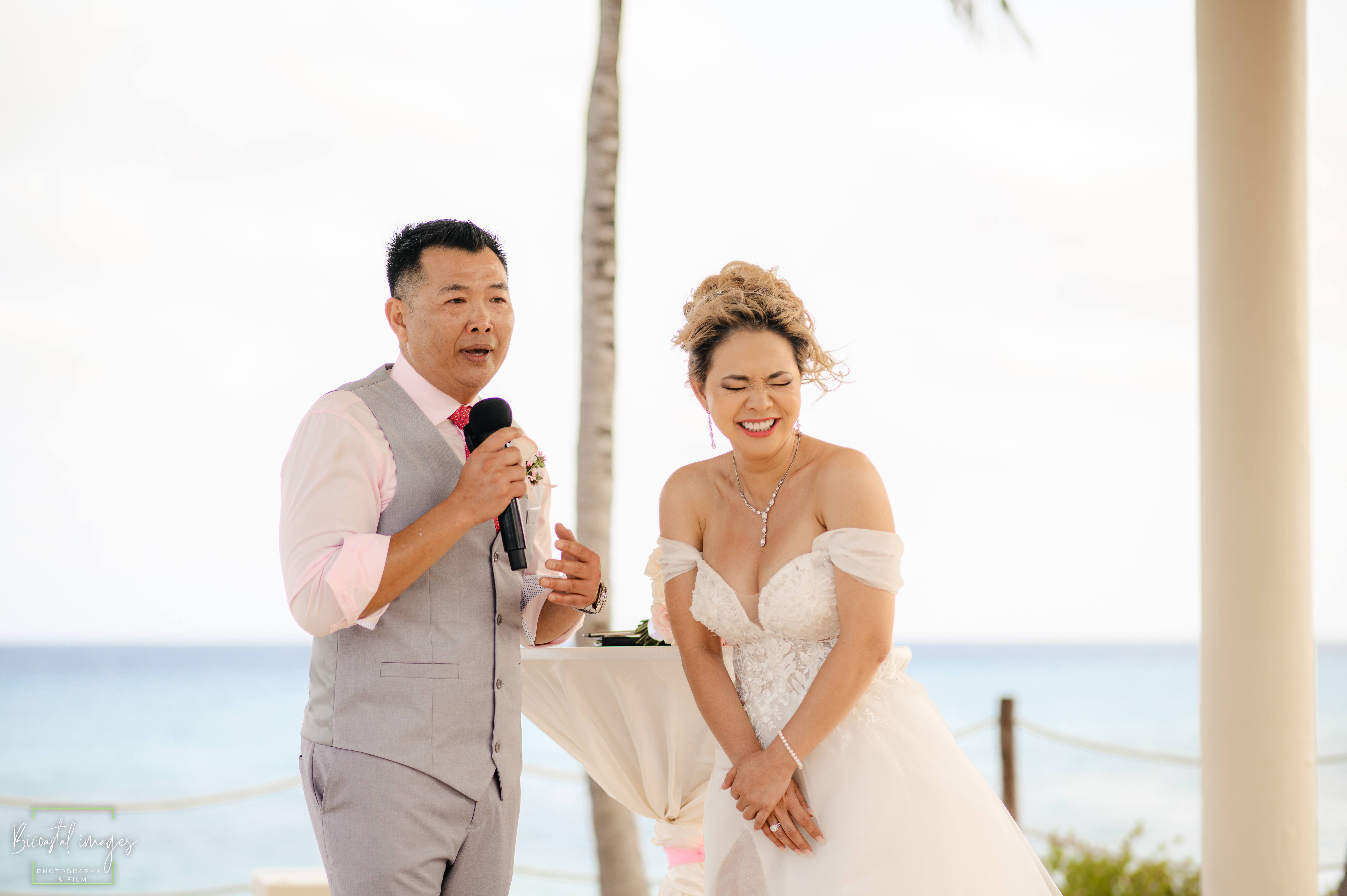 Groom speaking into microphone with bride laughing, ocean backdrop