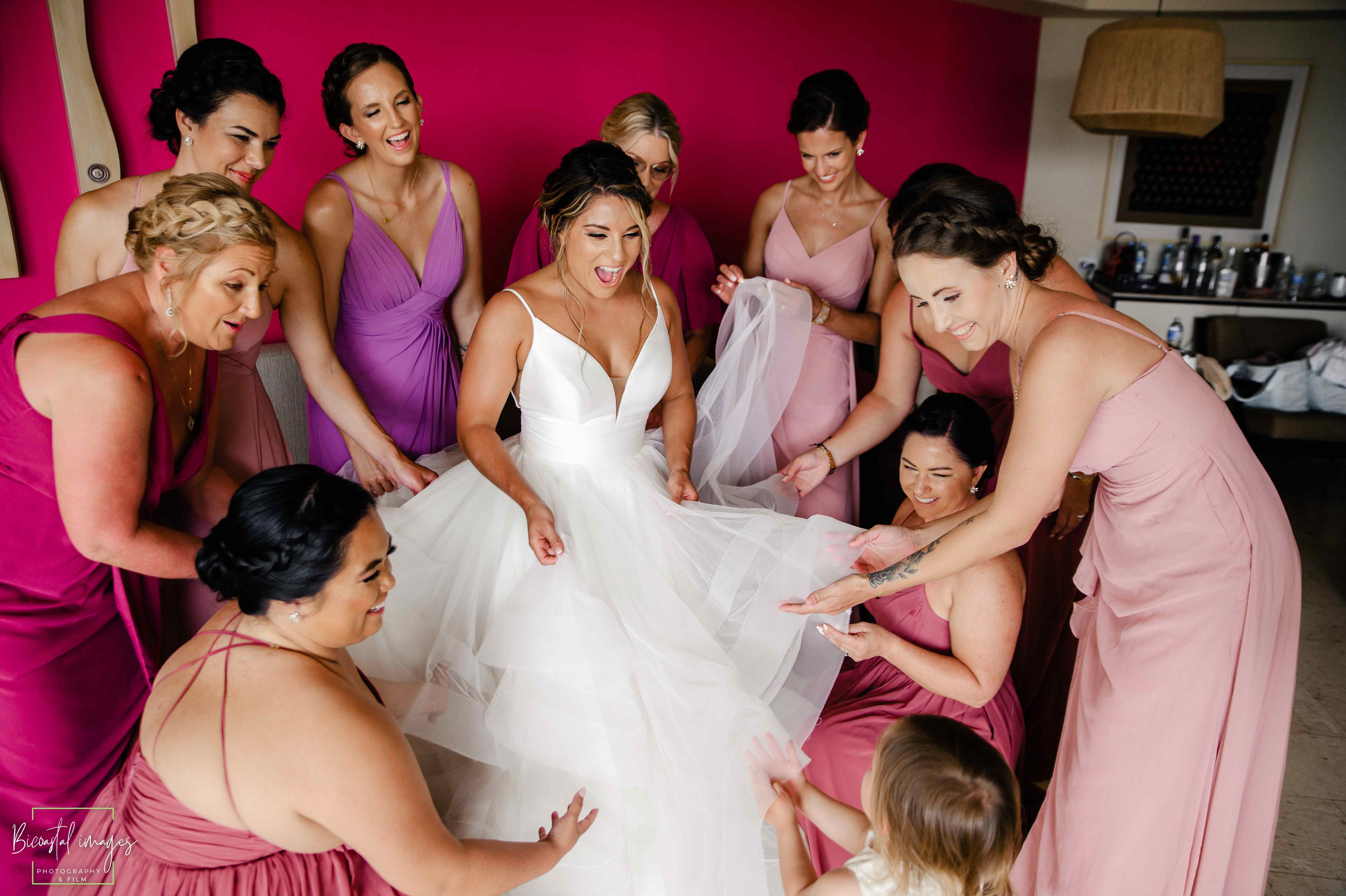 Bride in elegant white dress surrounded by bridesmaids in pink and burgundy, sharing intimate moment during destination wedding getting ready session in Riviera Maya