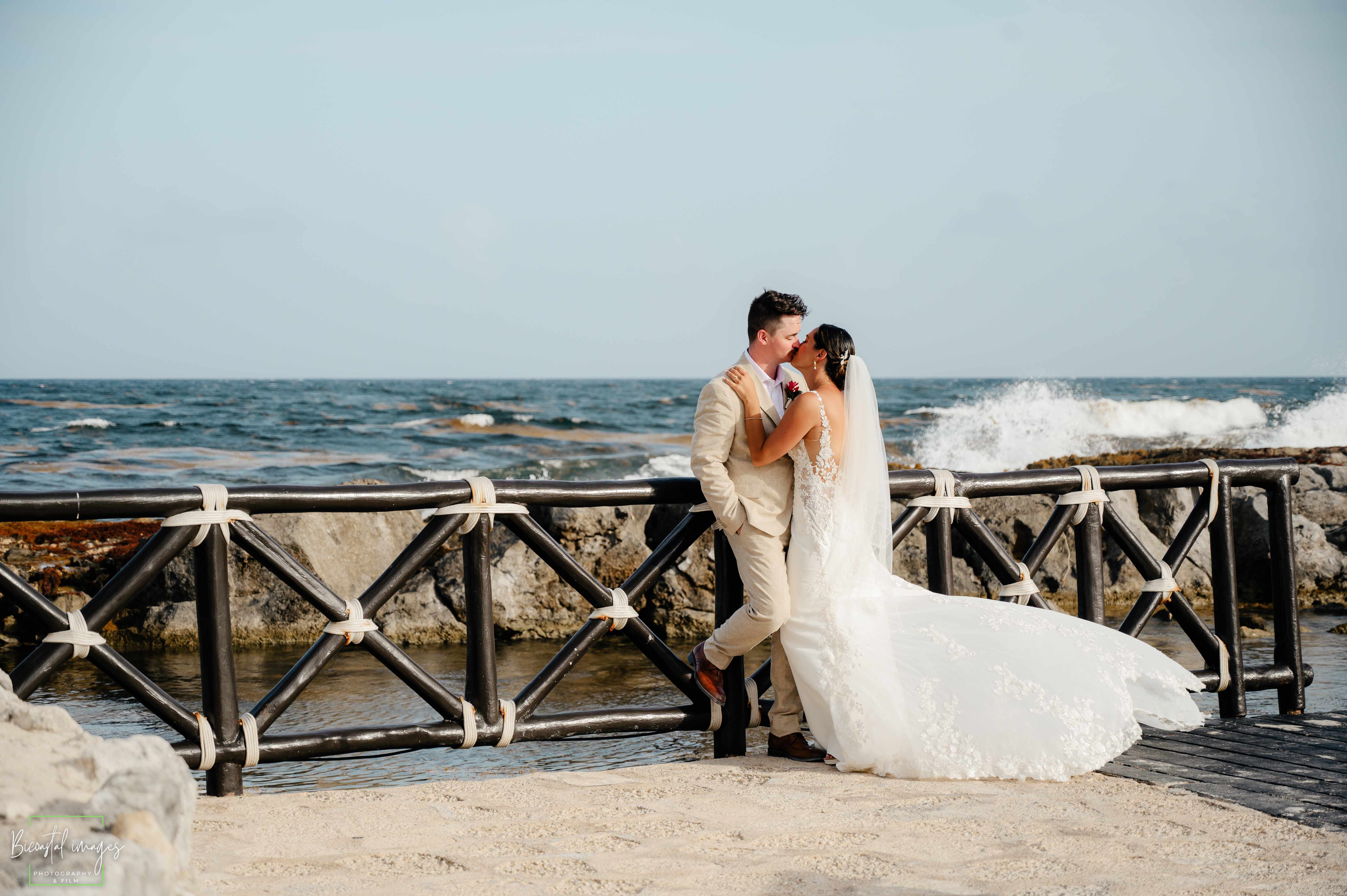 Bride and groom kissing on a pier by the rocky coastline with crashing waves at Hard Rock Riviera Maya destination wedding photographed by Bicoastal Images