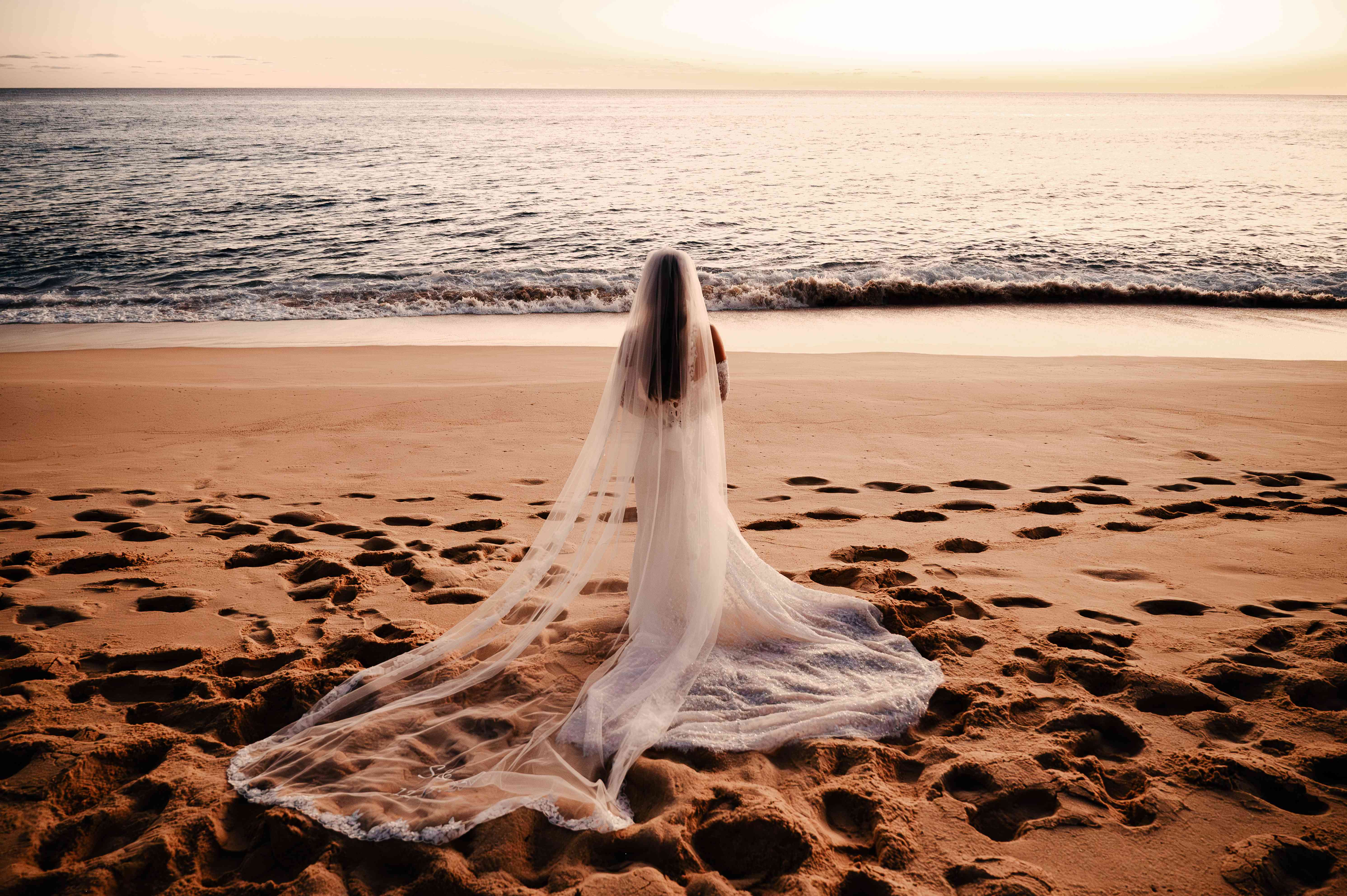 Bride with long flowing veil facing the ocean at golden hour on the beach at a Hard Rock resort in Mexico photographed by Bicoastal Images