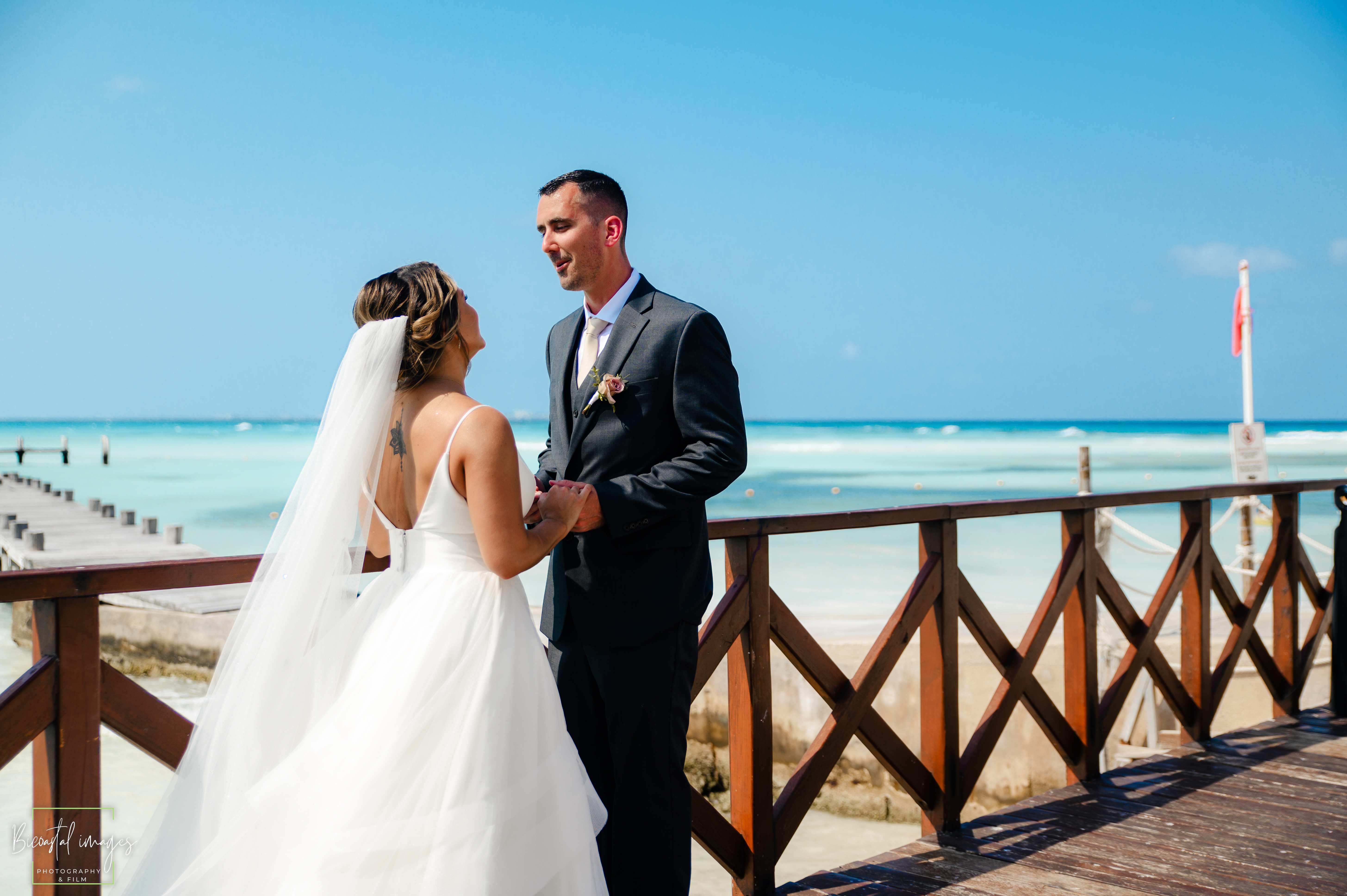 Bride and groom holding hands on a wooden pier over the turquoise Caribbean water at Hyatt Ziva Cancun destination wedding photographed by Bicoastal Images
